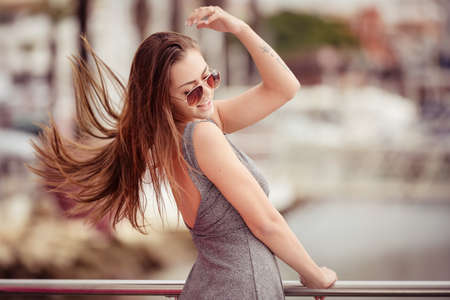 Portrait of beautiful young excited woman in sunglasses turning beside railing. Pretty girl in grey dress on blurred cityscape background.の写真素材