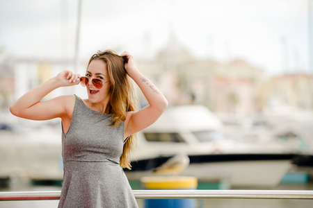 Portrait of beautiful young lady in sunglasses standing beside railing. Charming girl in grey dress on blurred embankment background.の写真素材
