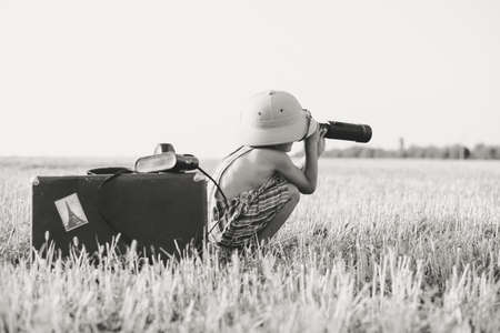 Picture of boy wearing romper looking in spyglass in country field. Black and white image of little explorer with camerabag and old suitcase on blurred countryside background.の写真素材