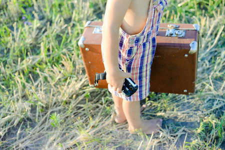 Picture of little boy with old suitcase and shock-proof mobile phone. Closeup of kid walking barefoot on sunny summer grass countryside background.の写真素材