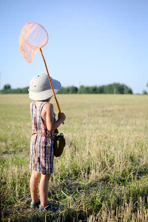 Picture of little boy with insect-net standing on field. Back view of kid wearing pith helmet on sunny summer countryside background.の写真素材