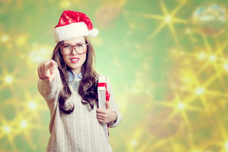 Portrait of beautiful young lady in red Santa Claus hat pointing finger and holding present box. Pretty girl in amazing glasses looking at camera on abstract festive design backgroundの写真素材