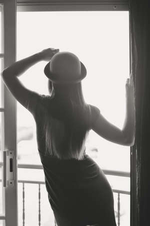 Black and white back view photo of amazing long haired female wearing dress and hat standing in bedroom doorwayの写真素材