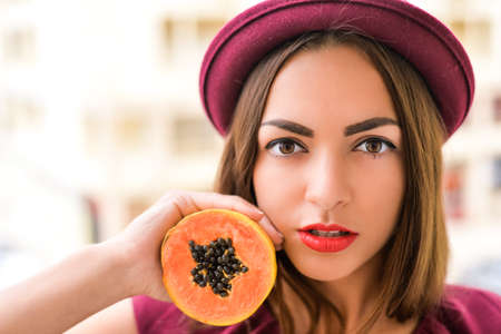 Portrait of elegant female with perfect makeup wearing red felt hat holding half of papaya fruit next to her faceの写真素材