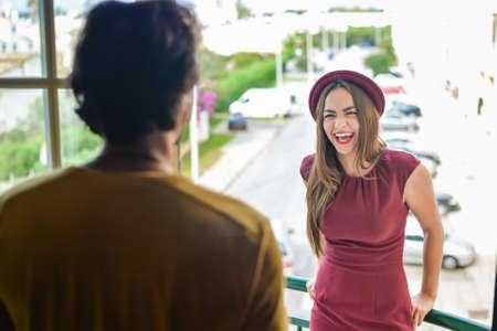 Photo of laughing young woman in red felt hat holding handrails on balcony looking at her man standing in doorwayの写真素材