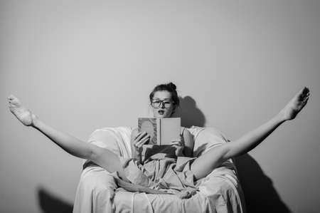 Picture of amazing beautiful young lady wearing glasses and holding a book relaxing split legs in chair. Black and white photographyの写真素材