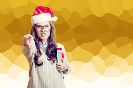 Portrait of beautiful young lady in red Santa Claus hat pointing finger and holding present box. Pretty girl in amazing glasses looking at camera on abstract festive design backgroundの写真素材