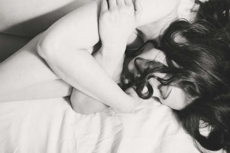 Closeup monochrome photo of lying on white bedsheet lady with her eyes closed embracing her kneesの写真素材