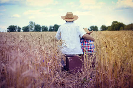 Picture of man and boy sitting on old suitcase in wheat farm field. Backview of happy family resting on summer countryside background.の写真素材
