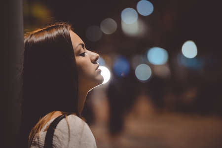 Closeup side view portrait of young sad thoughtful woman leaning against street lamp at night on bokeh copy space backgroundの写真素材