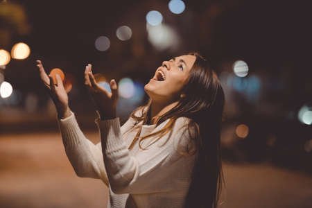 Closeup portrait of young lady in white sweater holding head and shouting in despair at night on street backgroundの写真素材