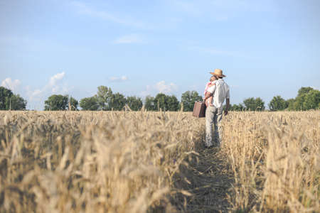 Picture of wondering father hugging baby and holding old suitcase in wheat field on sunny day. Backview of happy family standing on summer rural countryside road background.の写真素材