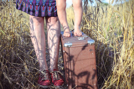 Picture of young woman and baby standing on old suitcase barefoot in wheat field. Closeup of happy family resting on summer countryside background.の写真素材