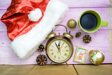 Picture of retro alarm clock with pine cones and Christmas ornaments. Santa hat and mug of coffee on pink plank table indoor background.の写真素材