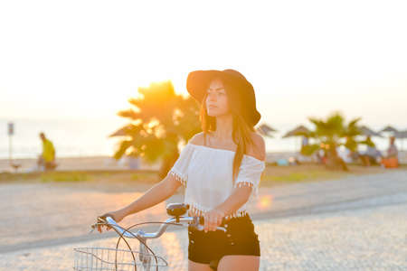 Lens flare photo of amazing lady wearing black floppy hat biking in sun flare on seaside backgroundの写真素材