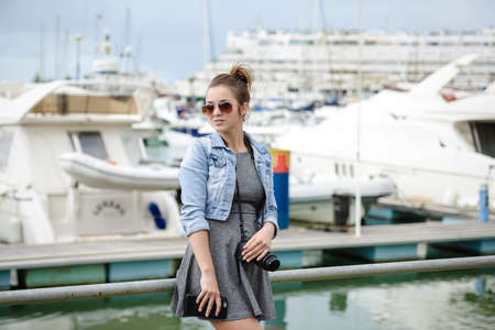 Picture of beautiful young lady holding camera standing in harbour. Backview of elegant photographer shooting yachts on summer sunny outdoor background.の写真素材