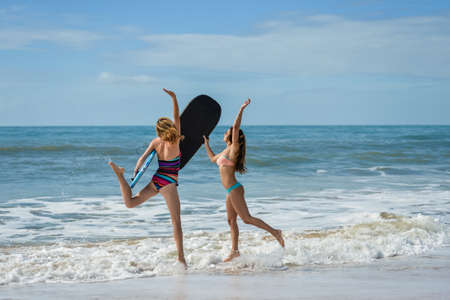 Healthy athletic young surfer girl friends with sexy fit body in bikini holding bodyboards, running on sea beach. Summer vacation on exotic sunny blue sky outdoors background.の写真素材