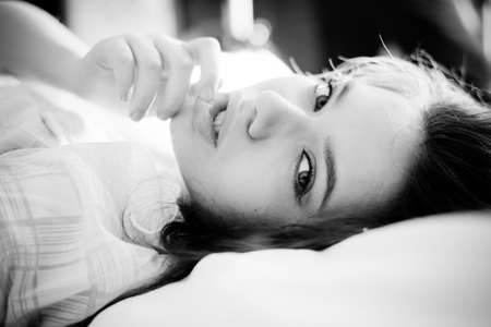 Black and white photography of beautiful young woman lying on bed touching her lips looking at cameraの写真素材