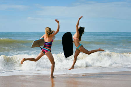 Healthy athletic young surfer girl friends with sexy fit body in bikini holding bodyboards, running...