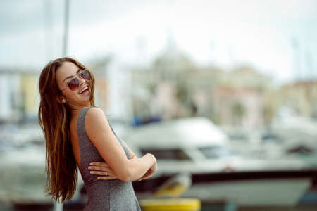 Outdoor portrait of young sexy tanned pretty woman standing on the pier near the sea marina. Back view of sensual girl in sunglassesの写真素材