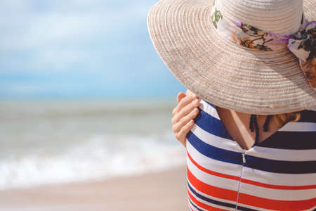 Back view of elegant beautiful female in dress and straw hat on the beach. Sunny blue sky background