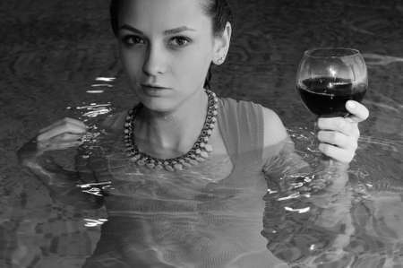 Black and white photography of young fashion model in evening dress standing in private swimming pool with glass of wine, closeup portraitの写真素材