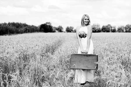 Black and white portrait of beautiful young lady in long dress with suitcase standing in the middle of wheat field outside backgroundの写真素材