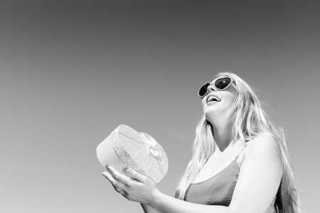 Black and white photography of young pretty blond lady in sunglasses holding heart shaped gift box over sky background copyspaceの写真素材