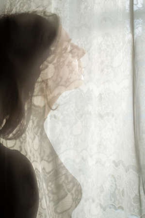 Beautiful young woman standing by the window between curtains on natural light backgroundの写真素材