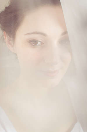 Beautiful young woman standing by the window between curtains on natural light backgroundの写真素材
