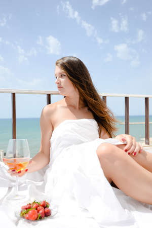 Beautiful woman luxury relaxing in bed enjoying glass of wine and strawberries on blue sky sea outdoors backgroundの写真素材
