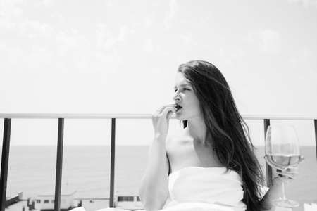 Black and white portrait of attractive woman relaxing in bed enjoying glass of wine and strawberries on sunny sky sea outdoors background. Enjoyment and luxury lifestyle.の写真素材