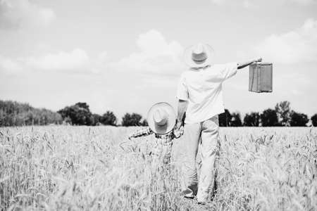 Black and white picture of father and son wearing straw hats holding old suitcase in wheat field. Happy vacation.の写真素材