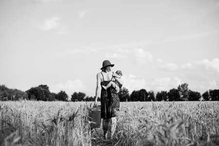 Black and white back view picture of woman with child standing in wheat field background. She carrying baby girl and suitcaseの写真素材