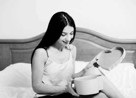 Happy Birthday and Valentine Day. Black and white picture of brunette girl excited with opening heart shaped gift box. Young pretty woman happy smiling on bedroom indoor background.の写真素材