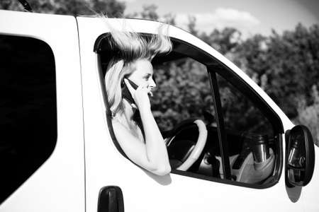 Black and white picture of emotional blond girl speaking phone and looking from car window. Young woman excited while talking with someone on sunny countryside background.の写真素材