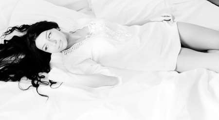 Black and white picture of young woman in lace nightclothes lying on bed sheet. Sexy lady with long tangled hair on blurred indoor background.の写真素材