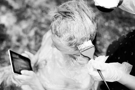 Black and white picture of hairdresser coloring hair using brush while client reading smartphone. Closeup of hand in glove and client's headの写真素材