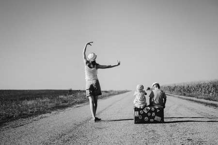 Black and white picture of young woman standing in ballet pose and two children sitting on old suitcase on country road. Back view of family traveling on sunny outdoors background.の写真素材
