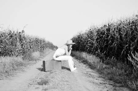 Black and white picture of woman wearing straw hat sitting on suitcase in remote country road. Sideview of female resting on sunny sky outdoors background.の写真素材