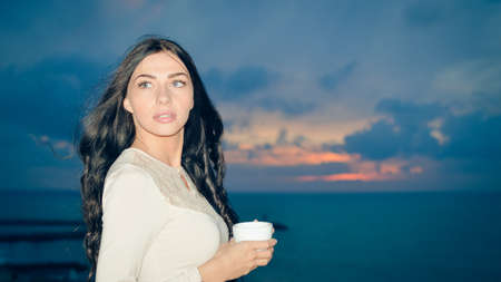 Portrait of elegant beautiful lady drinking a cup of hot drink, outdoors dawn background. Close upの写真素材
