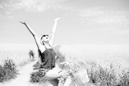 Black and white photography of joyful female sitting on bicycle lifting her arms with joy riding along rural road, field of wheat background outdoorsの写真素材