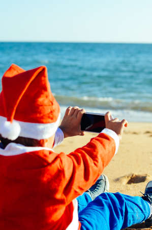 Back Side View of Joyful Child Santa Claus Using Smart Phone on Sunny Blue Sky Ocean Beach Outdoors Background. Mock up screen, taking photo, playing game on winter tropic holiday vacationの写真素材
