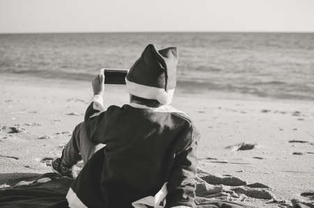 Back Side View of Joyful Child Santa Claus Using Smart Phone on Sunny Sky Ocean Beach Outdoors Background. Mock up screen, taking photo, playing game on winter tropic holiday vacation. Black and white pictureの写真素材
