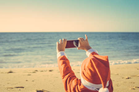 Back Side View of Joyful Child Santa Claus Using Smart Phone on Sunny Blue Sky Ocean Beach Outdoors Background. Mock up screen, taking photo, playing game on winter tropic holiday vacationの写真素材