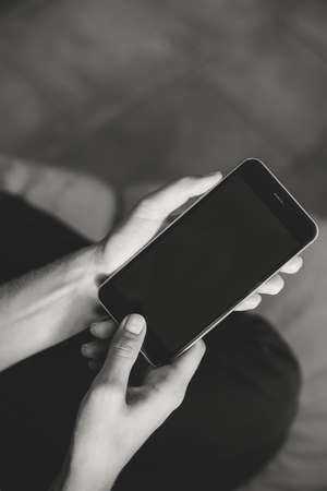 Black and white closeup on mobile phone in hand of young woman sitting on couch. Top side view mock up indoors backgroundの写真素材
