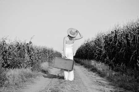 Black and white picture of woman wearing straw hat holding suitcase in corn field. Back view of happy joyful female on the road outdoors backgroundの写真素材