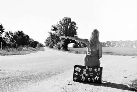 Black and white back view picture of woman sitting on suitcase on countryside road. Backview of female in dress hitchhiking car on sunny outdoors background.の写真素材