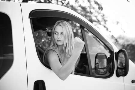 Black and white picture of angry blond girl in car showing fist on roadside. Young woman looking nervous after car incident on summer countryside background.の写真素材