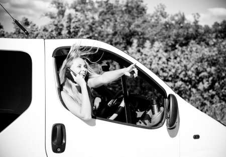 Black and white picture of blond young woman in car speaking on mobile phone and looking from car window. excited girl smiling while talking with someone on summer countryside background.の写真素材